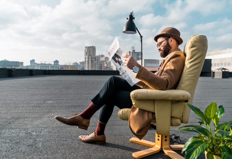 bearded businessman sitting in armchair and reading newspaper on roof